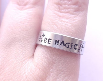 May include: A silver ring with the words "To Be Magic" engraved on it. The ring is worn on the middle finger of a hand with dark nail polish.