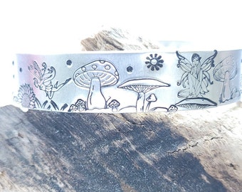 Puede incluir: Una pulsera de plata con un diseño grabado de hadas, setas y estrellas. La pulsera está sobre un trozo de madera a la deriva.