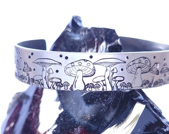 Puede incluir: Un brazalete plateado con un diseño grabado de setas. El brazalete abierto descansa sobre una superficie oscura y texturizada. El diseño de setas presenta setas de diferentes tamaños y formas. El brazalete es de metal plateado.