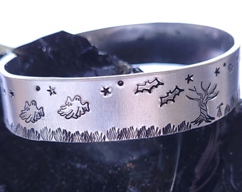 Puede incluir: Un brazalete de plata con un diseño de temática de Halloween. El brazalete presenta estrellas, fantasmas, murciélagos, acebo y un árbol.