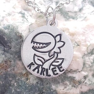 May include: A silver-colored pendant necklace featuring a stylized plant design with the name "KARLEE" engraved below. The pendant hangs from a delicate chain and rests on a textured, light green and white stone surface.
