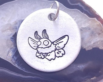 May include: A silver charm featuring a detailed engraving of a moth with large eyes and feathery wings. The charm is resting on a slice of agate with brown and white bands.