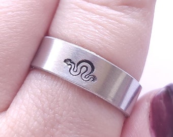 May include: A silver-colored ring with a flat band. The ring features a small, engraved snake design. The ring is shown on a finger with dark red nail polish.