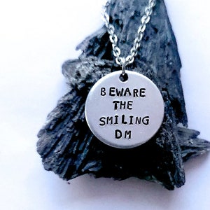 Puede incluir: Un collar de plata con un colgante redondo que dice "Beware the Smiling DM".