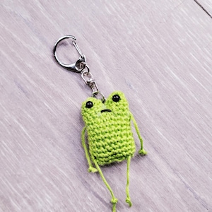 May include: A handmade green crochet frog keychain with black eyes and dangling legs. The frog is attached to a silver-colored metal clasp. The background is a light gray wooden surface.