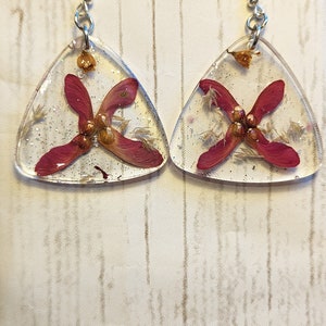 Puede incluir: Un par de pendientes de forma triangular con una base de resina transparente y flores rojas prensadas en su interior. Los pendientes tienen ganchos plateados.