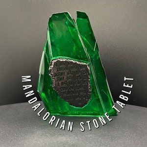 May include: A translucent green stone tablet with a dark, textured inset featuring an inscription. The words "MANDALORIAN STONE TABLET" are arched around the base of the stone.