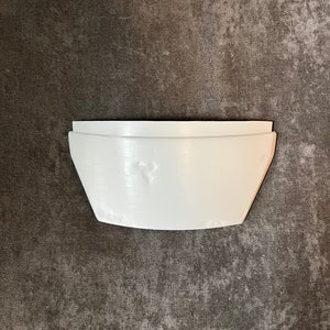 May include: A white, geometric-shaped planter with a curved bottom and straight sides. The planter is made of a solid material and has a smooth, matte finish. The design is modern and minimalist, suitable for small plants.