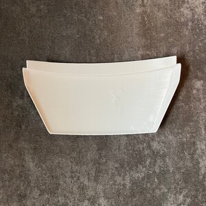 May include: A white, rectangular, curved-edge object with a trapezoidal shape. The object has a smooth surface and appears to be made of plastic or a similar material. The item is set against a dark gray, textured background.