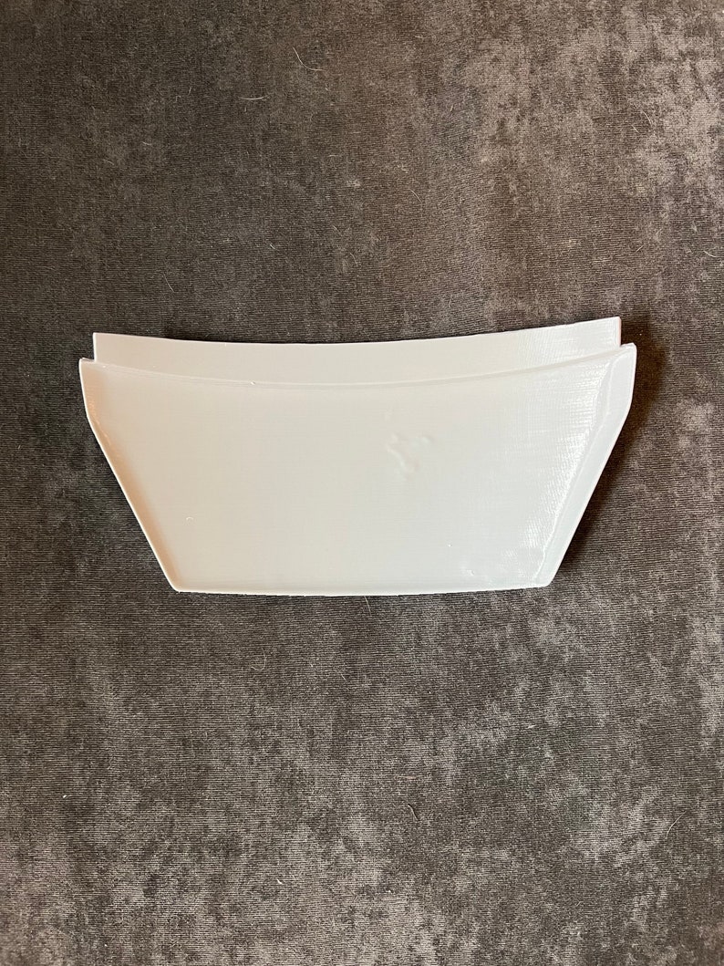 May include: A white, rectangular, curved-edge object with a trapezoidal shape. The object has a smooth surface and appears to be made of plastic or a similar material. The item is set against a dark gray, textured background.