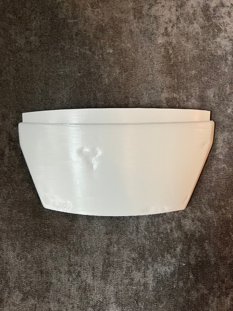 May include: A white, rectangular object with a curved top and angled sides. The object appears to be made of a solid material and is resting on a dark, textured surface. The shape suggests it could be a shelf or a similar storage component.