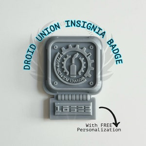 May include: A gray 3D printed Droid Union Insignia Badge with the text "Droid Union Insignia Badge" around the perimeter. The badge features a gear with a wrench and a small cylinder inside. The badge also has a rectangular area with the text "1A323". The text "With FREE Personalization" is below the badge.