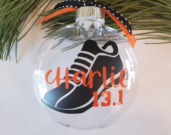 Puede incluir: Adorno de Navidad de vidrio transparente con una zapatilla de correr negra y el nombre "Charlie" y "13.1" en letras naranjas.