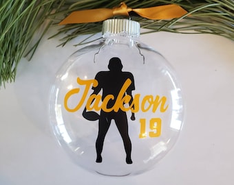 May include: Clear glass Christmas ornament with a black football player silhouette, the name "Jackson" in yellow script, and the number "19" in yellow. The ornament has a gold ribbon for hanging.