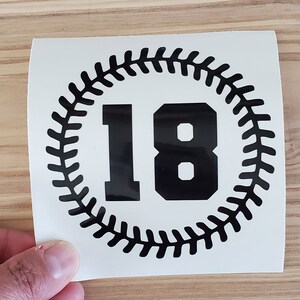 May include: A black and white sticker featuring the number 18 in a bold font, encircled by a baseball stitch design. The sticker is held against a light wood surface.