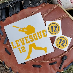 May include: White stickers with yellow baseball-themed graphics and text. One sticker features a batter silhouette and the name "LEVESQUE" with the number "12". Two smaller stickers display the number "12" in a blue circle with a yellow border. The stickers rest on a brown baseball glove.