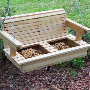 May include: A wooden bird feeder shaped like a swing bench with two compartments filled with birdseed. The feeder is hanging from a chain.