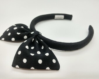 May include: Black headband with a white polka dot bow. The headband is made of fabric and is a simple, elegant accessory.