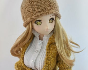 Peut inclure: Une poupée aux cheveux blonds portant un bonnet en tricot marron avec un détail de feuille verte, une chemise blanche boutonnée, un cardigan en tricot jaune et une jupe à carreaux.