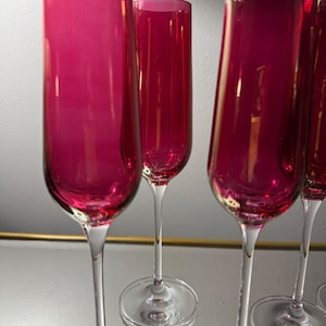 Könnte beinhalten: Eine Reihe von Champagnerflöten in Magenta. Die schlanken Gläser haben klare Stiele und Böden. Sie sind auf einer reflektierenden Oberfläche angeordnet, was eine elegante Ästhetik erzeugt. Ideal für festliche Anlässe.