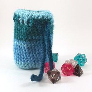 Puede incluir: Una bolsa de dados de crochet azul y verde con cierre de cordón. La bolsa está llena de dados de colores.
