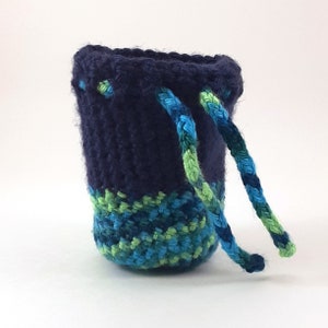 Puede incluir: Una pequeña bolsa de cordón de ganchillo con un cuerpo azul marino y un fondo estampado en verde y azul. La bolsa tiene dos cordones largos trenzados.