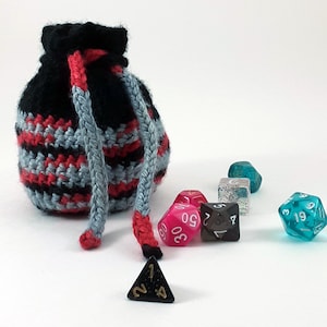 Puede incluir: Una bolsa de dados de crochet negra y gris con cierre de cordón. La bolsa está llena de dados de colores, incluyendo dados rosas, azules y transparentes con números blancos.