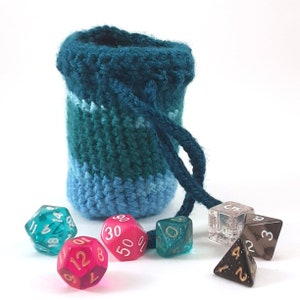 Puede incluir: Una bolsa de dados de crochet azul turquesa con cierre de cordón. La bolsa está llena de seis dados de colores, incluyendo un dado azul, verde, rosa y negro.