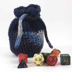 Puede incluir: Una bolsa de dados de crochet azul con cierre de cordón, llena de seis dados coloridos. Los dados están numerados del 1 al 6 y tienen una variedad de colores, incluyendo rojo, verde, amarillo y naranja.