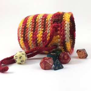 Puede incluir: Una bolsa de dados de crochet con cierre de cordón en tonos rojos, naranjas, amarillos y marrones. La bolsa está llena de dados de varios colores, incluyendo rojo, verde y negro.