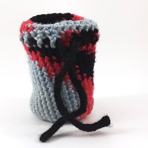 Puede incluir: Una pequeña bolsa de cordón de ganchillo con un patrón de color rojo, negro y gris. La bolsa tiene un cordón negro.
