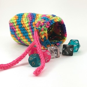 Puede incluir: Una bolsa de dados de crochet colorida con un cordón de ajuste rosa. La bolsa está llena de varios dados, incluyendo un d6 turquesa, un d6 negro, un d20 rosa y un d6 transparente.