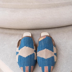 May include: A pair of blue and beige patterned slippers with brown trim. The slippers have a geometric design with a beige central pattern and vertical stripes at the toe. The background is a neutral color.