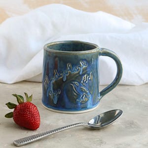 May include: A handmade ceramic mug with a blue glaze, featuring the text "Muddybanks" and a bird design. A fresh red strawberry and a silver spoon are next to the mug. The mug has a curved handle and a white base.