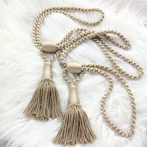 May include: Two beige curtain tiebacks with braided cords and decorative tassels. The tassels are made of twisted strands, and the cords have a loop at the end. The tiebacks are designed to hold curtains open and add a decorative touch to a room.