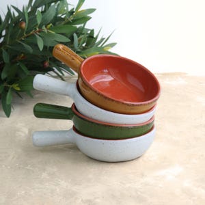 May include: A stack of four ceramic bowls with handles. The top bowl is orange with a tan rim and handle. The next bowl is white with a speckled design and a white handle. The third bowl is green with a white handle. The bottom bowl is white with a speckled design and a white handle.