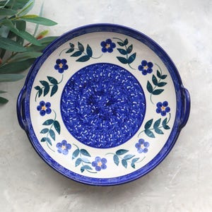 May include: Round ceramic serving platter with a blue floral design. The platter features a white rim with blue flowers and green leaves. The center of the platter is a textured blue pattern, and it has two handles.