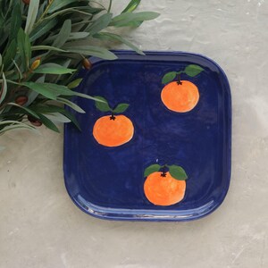 May include: A square, cobalt blue ceramic tray with rounded corners. The tray is decorated with three hand-painted orange citrus fruits, each with green leaves. Olive branches are in the upper left corner.