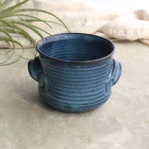 May include: A handcrafted, blue ceramic planter with a textured, ribbed design. The planter features two small handles and a dark brown rim. The interior is a darker shade of blue. Ideal for small plants, this planter measures approximately 6 inches in diameter and 5 inches tall.