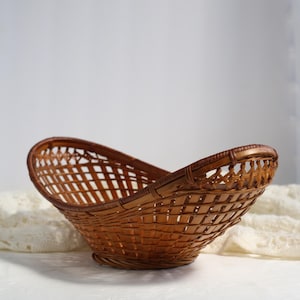 May include: A woven brown basket with a curved, boat-like shape. The basket is made of thin, interwoven strips of wood, creating a lattice pattern. The rim is reinforced with a thicker, darker brown material. The basket sits on a white surface.