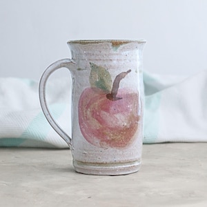 May include: A tall, off-white ceramic mug with a large, hand-painted apple design. The apple is primarily red with green and brown accents. The mug has a curved handle and a slightly flared rim, perfect for enjoying hot beverages.