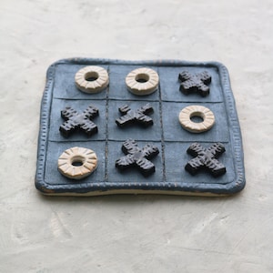 May include: A handcrafted tic-tac-toe game with a blue square board and nine game pieces. The game pieces are a mix of beige circles and dark brown X shapes. The board has a textured surface and a decorative border.