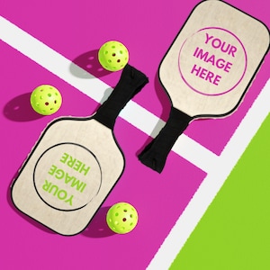 May include: Two pickleball paddles with black handles and text that reads "YOUR IMAGE HERE" are displayed on a pink and green court. Three yellow pickleballs with holes are scattered around the paddles.