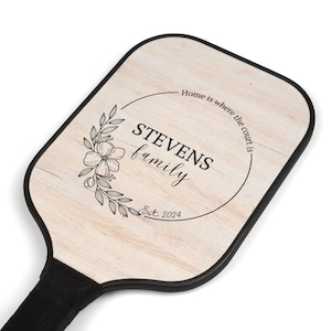 Könnte beinhalten: Pickleball-Schläger mit hellem Holzmaserungsdesign. Der Schläger hat einen schwarzen Rand und Griff. Die Vorderseite des Schlägers hat ein florales Kranzdesign mit dem Text "STEVENS family" und "Home is where the court is."