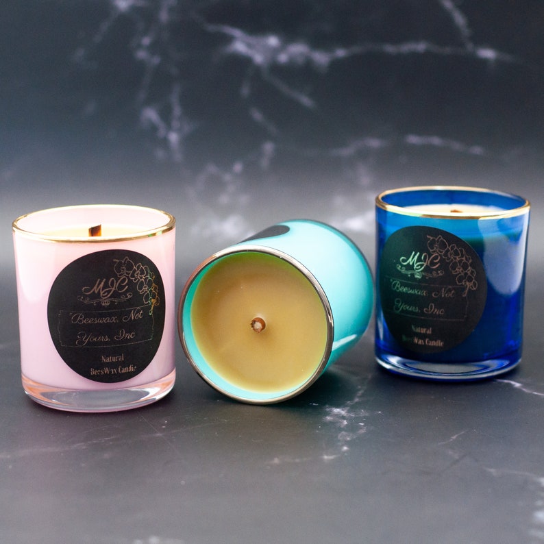 May include: Three glass jar candles with gold rims. One is light pink, one is turquoise, and one is deep blue. Each features a black circular label with the text MJC, Beeswax, Not Yours, Inc, and Natural Beeswax Candle. Each has a single wick.