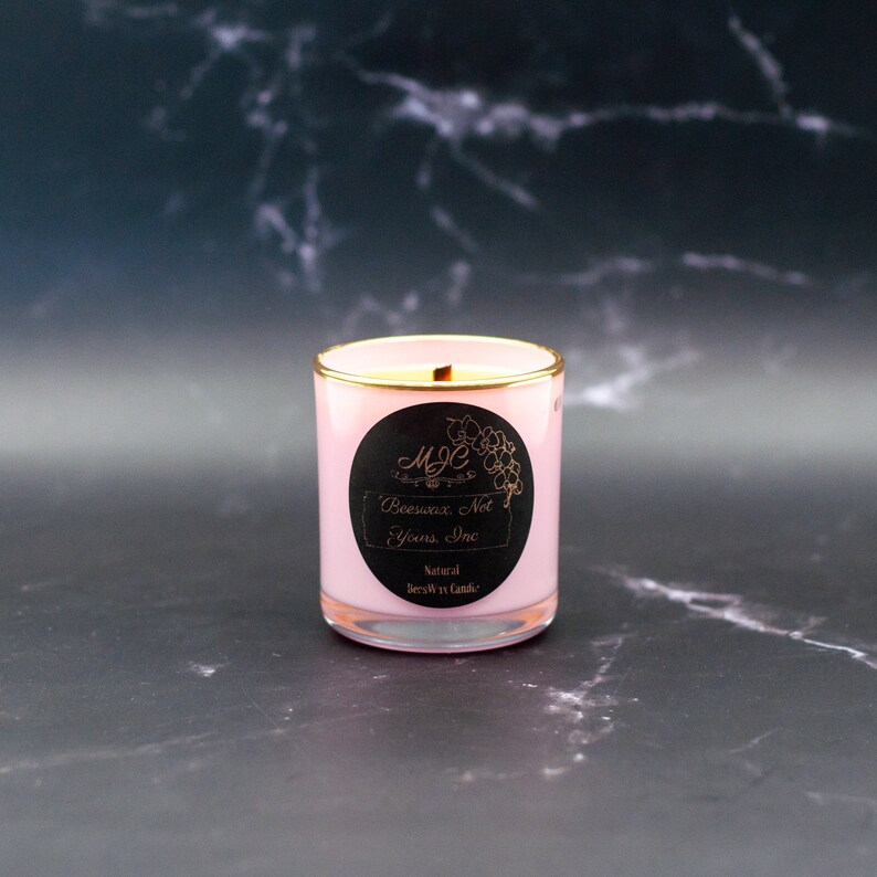 May include: A light pink candle in a glass jar with a gold-toned rim. A circular black label on the front features the text MJC, an orchid illustration, and the words Beeswax, Not Yours, Inc. along with the text Natural BeesWax Candle.