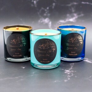 May include: Three glass candles in dark green, turquoise, and blue, each with a gold rim and a wooden wick. A circular black label on each features the text MJC, Beeswax, Not Yours, Inc, and Natural BeesWax Candle with an orchid illustration.