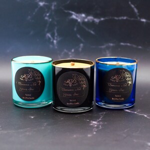 May include: Three glass candles in turquoise, black, and blue with gold rims. Each features a round black label with gold text reading MJC Beeswax, Not Yours, Inc and Natural BeesWax Candle, plus a floral graphic. Each candle has a visible wick.