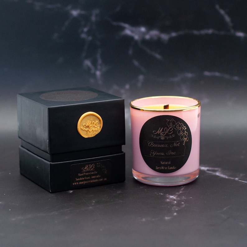 May include: A light pink candle in a glass jar with a gold rim and a black circular label reading MJC Beeswax, Not Yours, Inc. Natural Beeswax Candle. Beside it sits a black square box with a gold wax seal featuring the letters MJC.