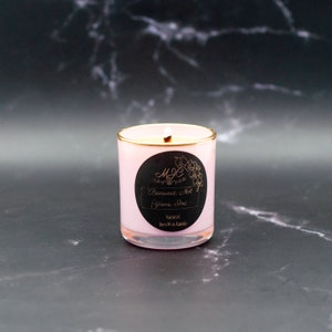 May include: A light pink candle in a glass jar with a gold rim. A black circular label features the text MJC, Beeswax, Not Yours, Inc, and Natural BeesWax Candle. The candle has a single lit wick, and the wax inside is a pale cream color.